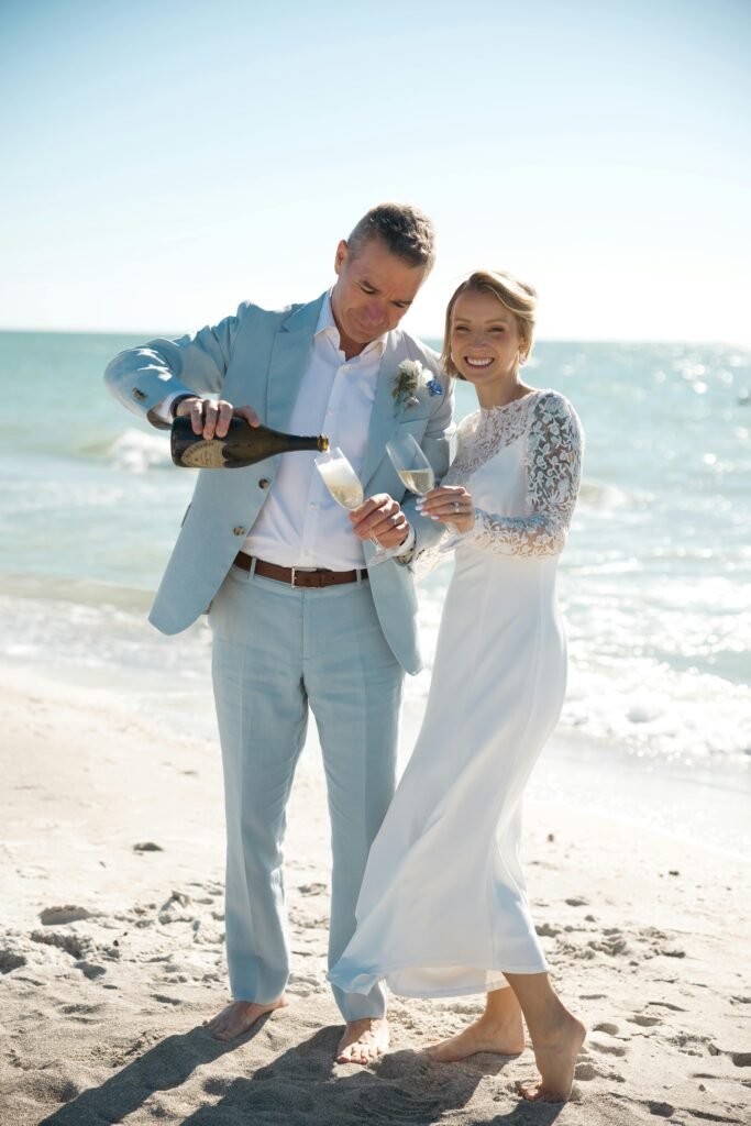 A joyful bride and groom celebrate with champagne by the ocean under clear skies.